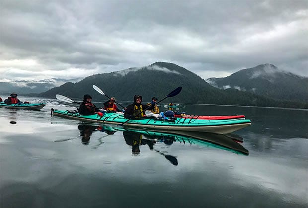 Imaeg of 4 people in 2 kayaks on still water with mountains in the distance