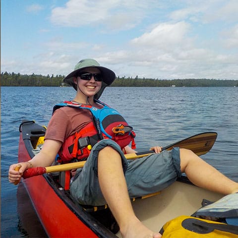 Image of aperson in a hat and lifejacket, sitting in the back of a canoe