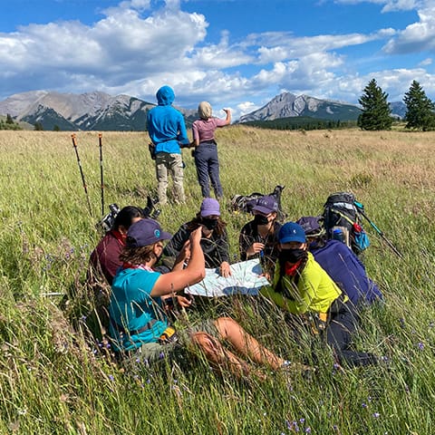 Image of a group of of participants sitting in a field looking at a map. Two other are standing behind them pointing at the mountains