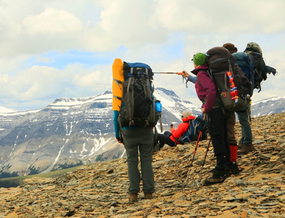Group of four hikers with large backpacks and walking sticks standing on a rocky slope pointing at a mountain peak