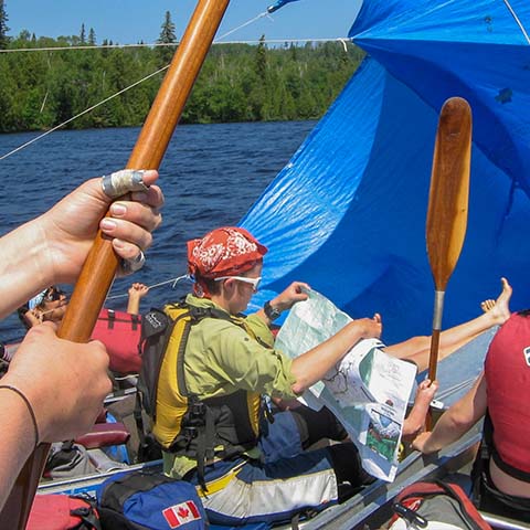 Image of a person holding a map in a canoe. Other are making a sail out of paddles and a tarp