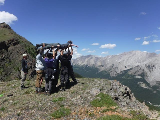 Group of people lifting one person on their stomach, near the edge of a cliff, mountains in distance