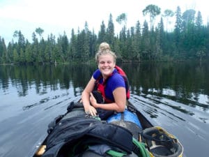 Person smiling in the front of a canoe on a still lake. Trees on the shore in the distance.