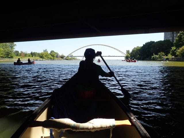 Silouette of a person canoeing under a bridge with a suspension bridge in front of them