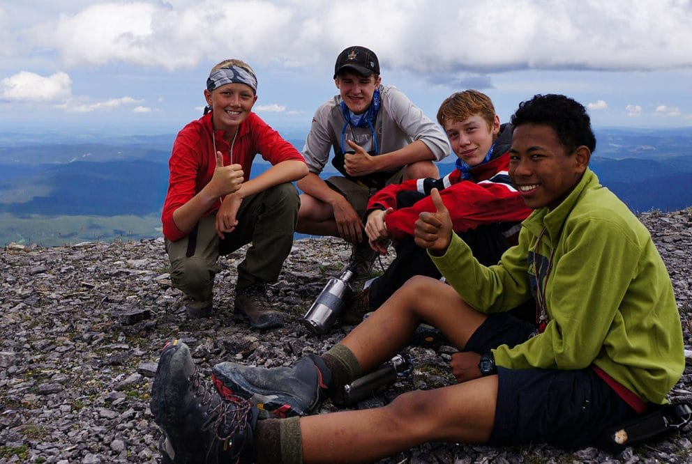 Image of four teens sitting on the top of mountains showing thumbs up