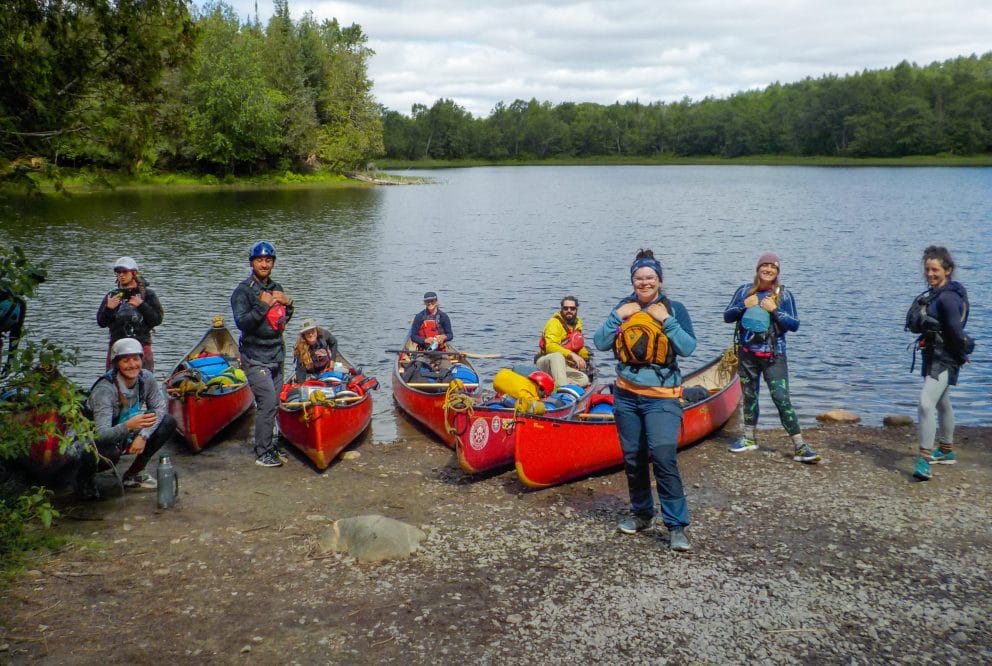 Image of a group of instructors at Ontario staff training. The group is on the shore of the lake with a handful of canoes.