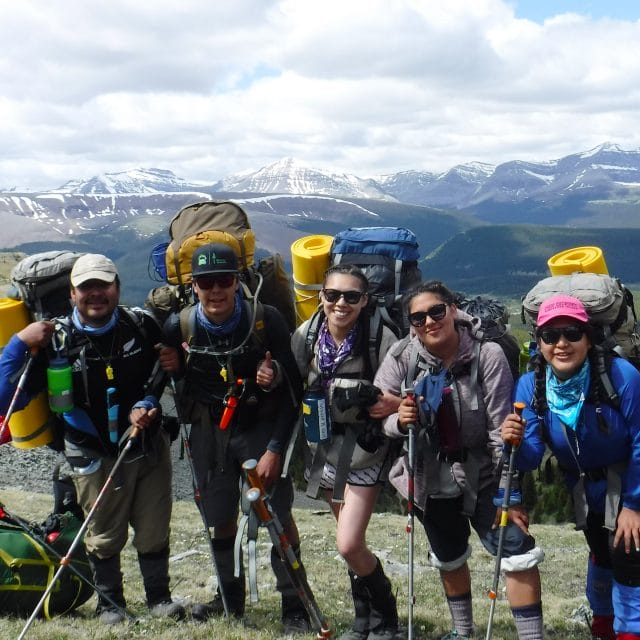 Image of 5 people with large hiking backpacks with mountain tops behind them
