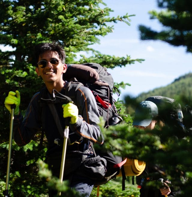 Image of Aryan standing in front of tree covered mountain. He is holding 2 hiking poles.