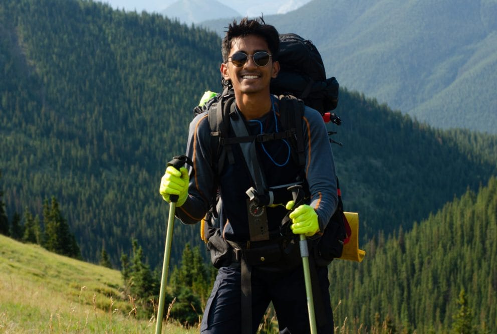 Image of Aryan standing in front of tree covered mountain. He is holding 2 hiking poles.