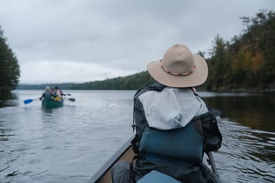 Image of someone in a hat canoeing on a lake. Another canoe with 2 paddlers is in the distance.