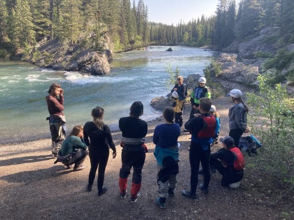 group of staff training instructors standing in a semi-circle by a river in the Rocky Mountains listening to speaker