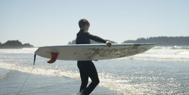 Image of a teen in a wetsuit, carrying a surfboard